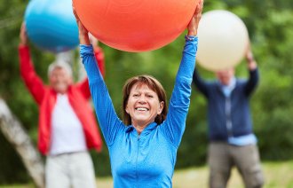 Bildet viser en smilende eldre kvinne i treningstøy som holder en stor ball over hodet.