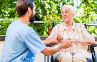 Bildet viser en pleier som holder hendene til en eldre kvinne som sitter i rullestol