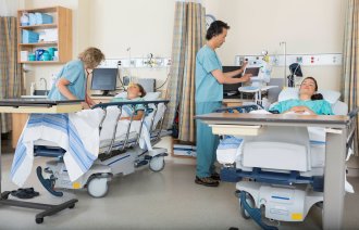 Bildet viser to pasienter og to sykepleiere som står ved sengene deres på postoperativen.