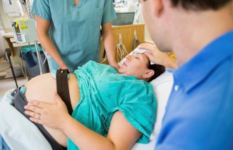 Bildet viser en kvinne som holder på å føde. Sykepleierne og legen måler fosterlyden, mens faren tørker pannen til kvinnen.