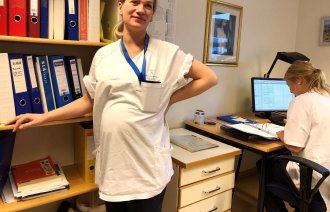 Høygravid sykepleier tar seg til ryggen og lener seg til bokhylle. Kollega sitter foran skjerm ved sine av henne.