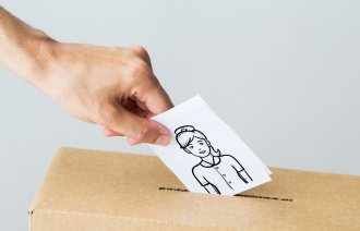 Bildet viser en hånd som slipper en stemmeseddel ned i en papireske. På stemmeseddelen er det bilde av en sykepleier.