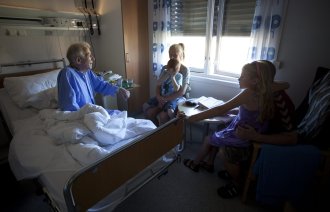 Bildet viser en syk, eldre mann i en sykehusseng. Han har besøk av barn og barnebarn.