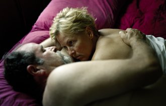 Et voksent par ligger i sengen halvveis under dyna og ser på hverandre