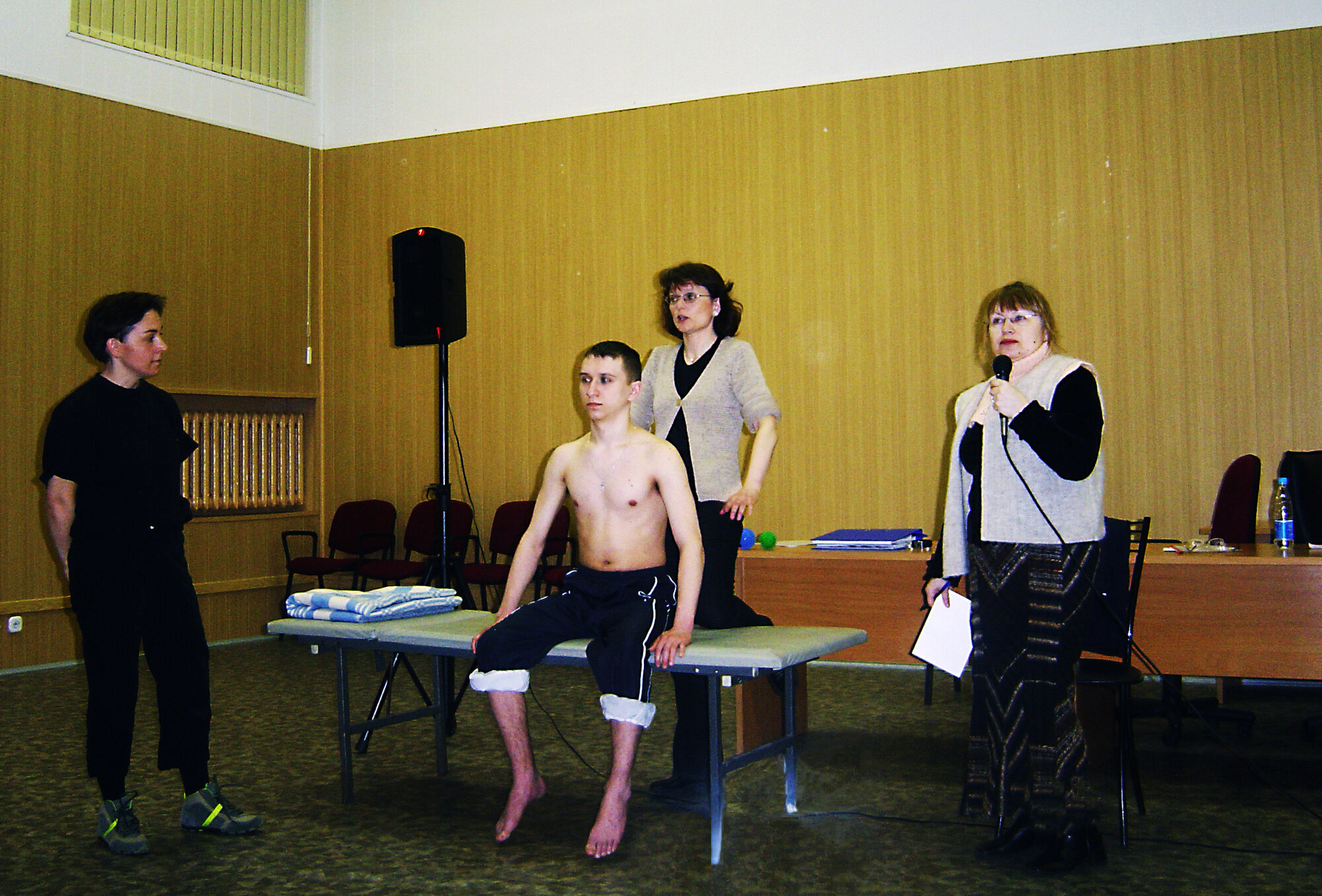 Fysioterapeuter demonstrerer foran et russisk publikum. Bildet viser to norske fysioterapeuter som demonstrerer foran et russisk publikum. En mannlig pasient sitter på en seng, mens en tolk oversetter.