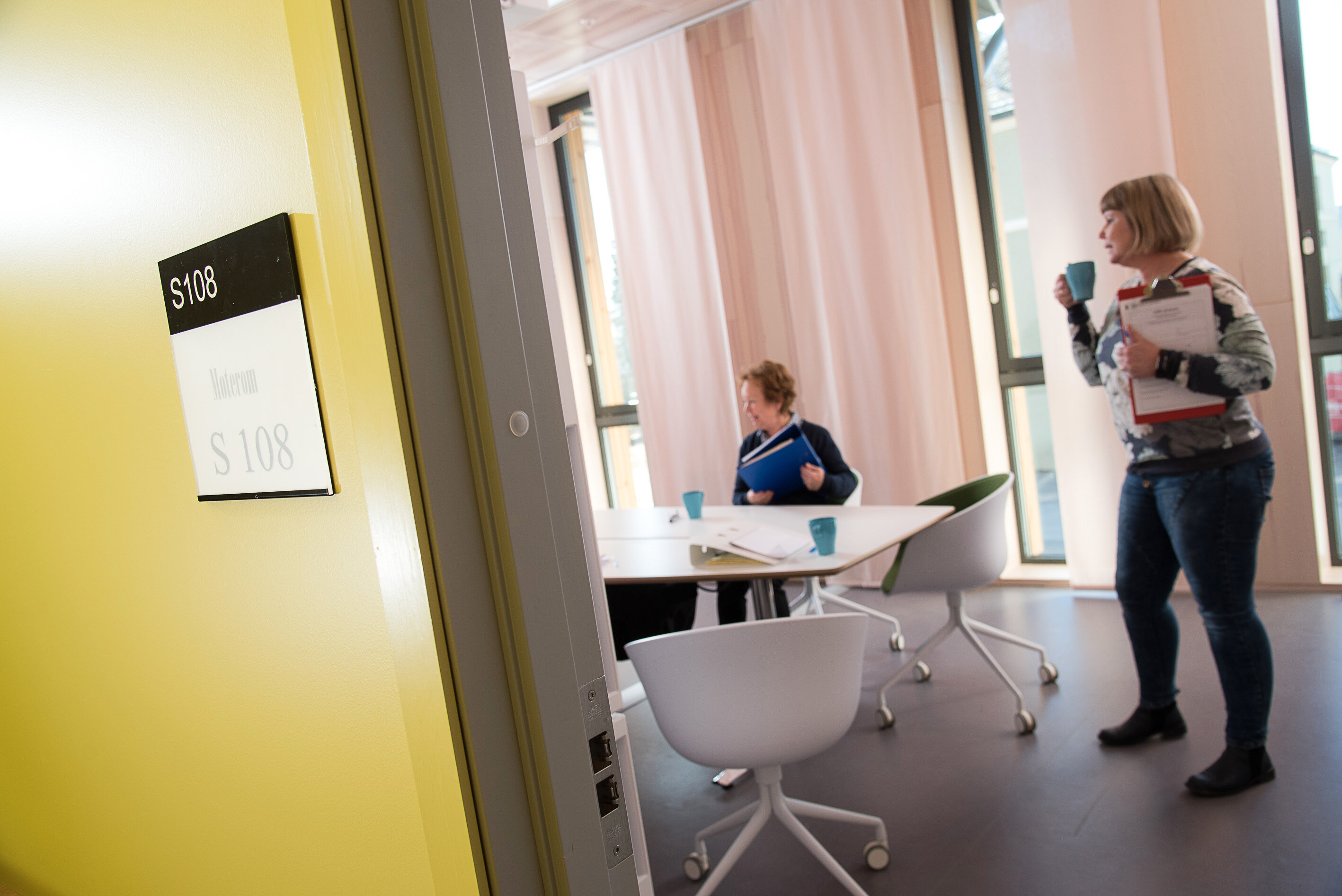 Samarbeidsmøte Bildet viser døråpningen til et møterom, der det er to personer. Den ene sitter ved bordet, og den andre står ved siden av og drikker en kopp kaffe.