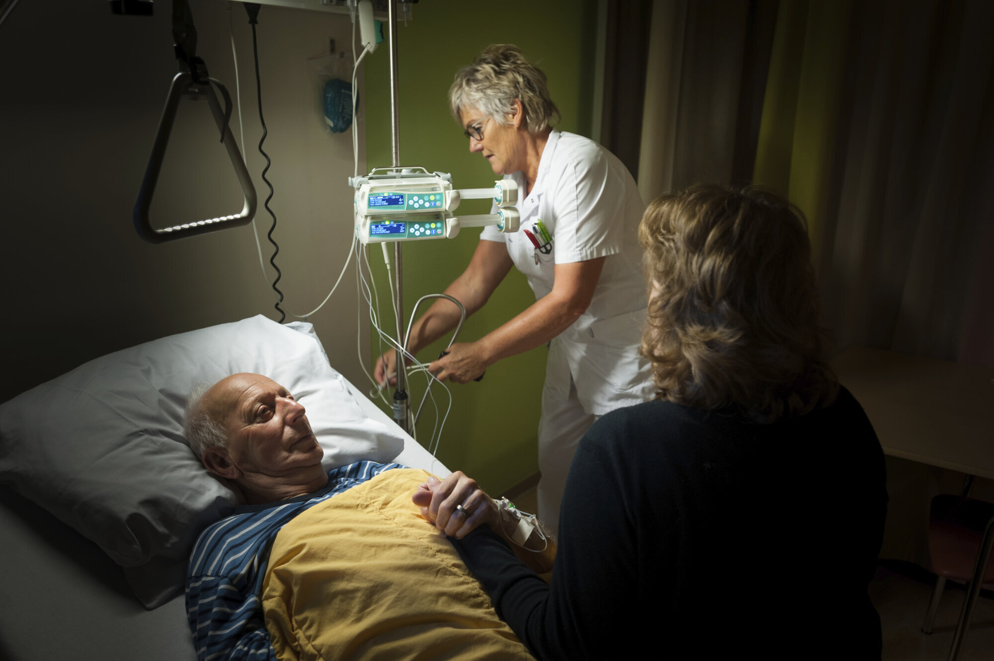 Bildet viser en mann i sengen på palliativ avdeling. En sykepleier sjekker noen slanger, mens en pårørende kvinne sitter og holder mannen i hånda.