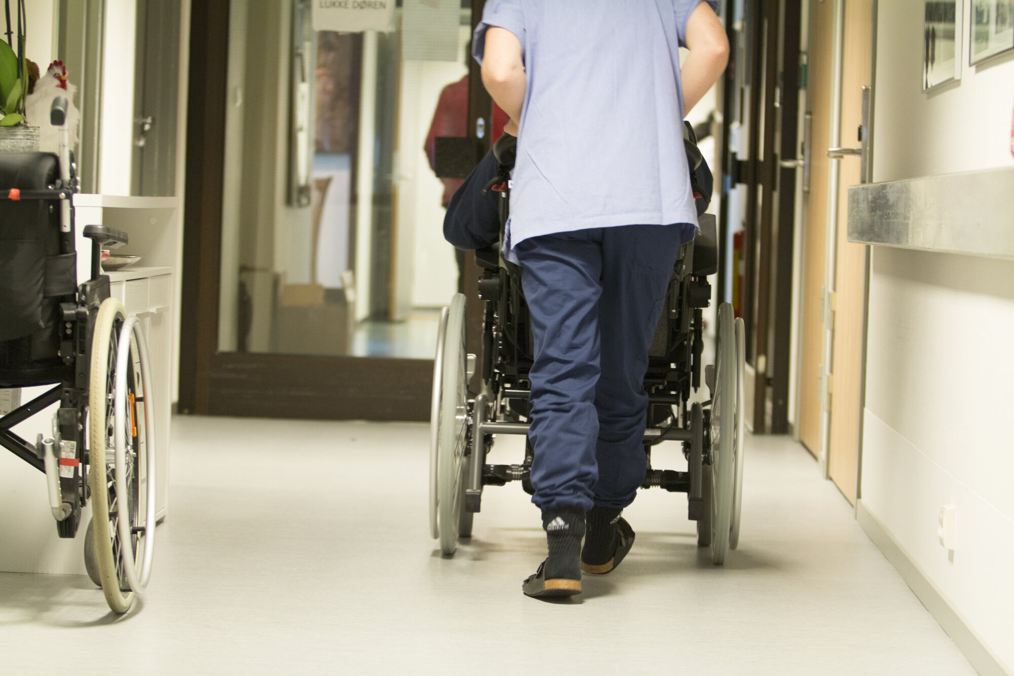 Bildet viser en sykepleier som triller en eldre pasient i rullestol.