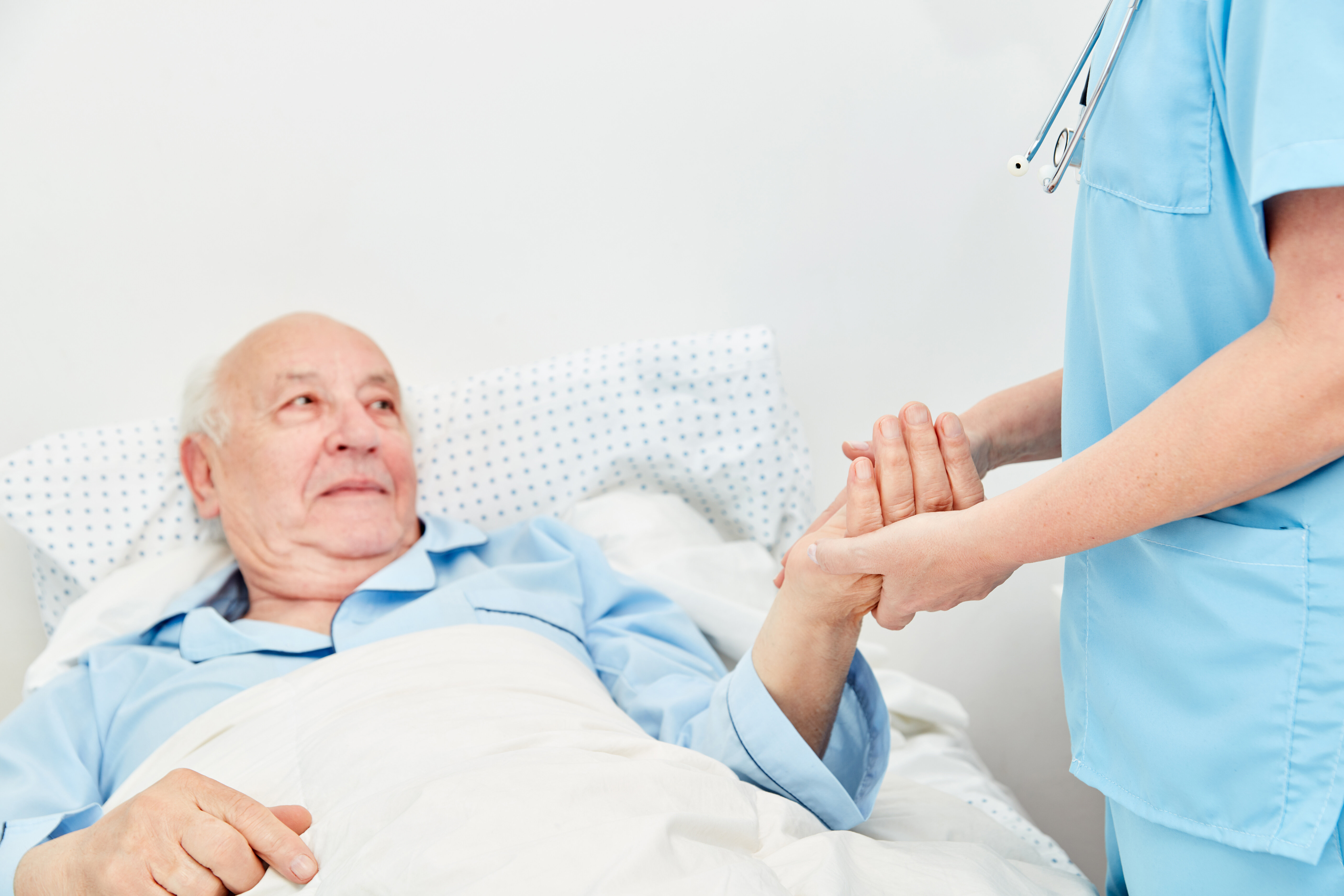 Sykepleier og eldre mann på sykehjem Bildet viser en eldre mann som ligger i senga. En sykepleier står ved siden av og holder ham i hånda