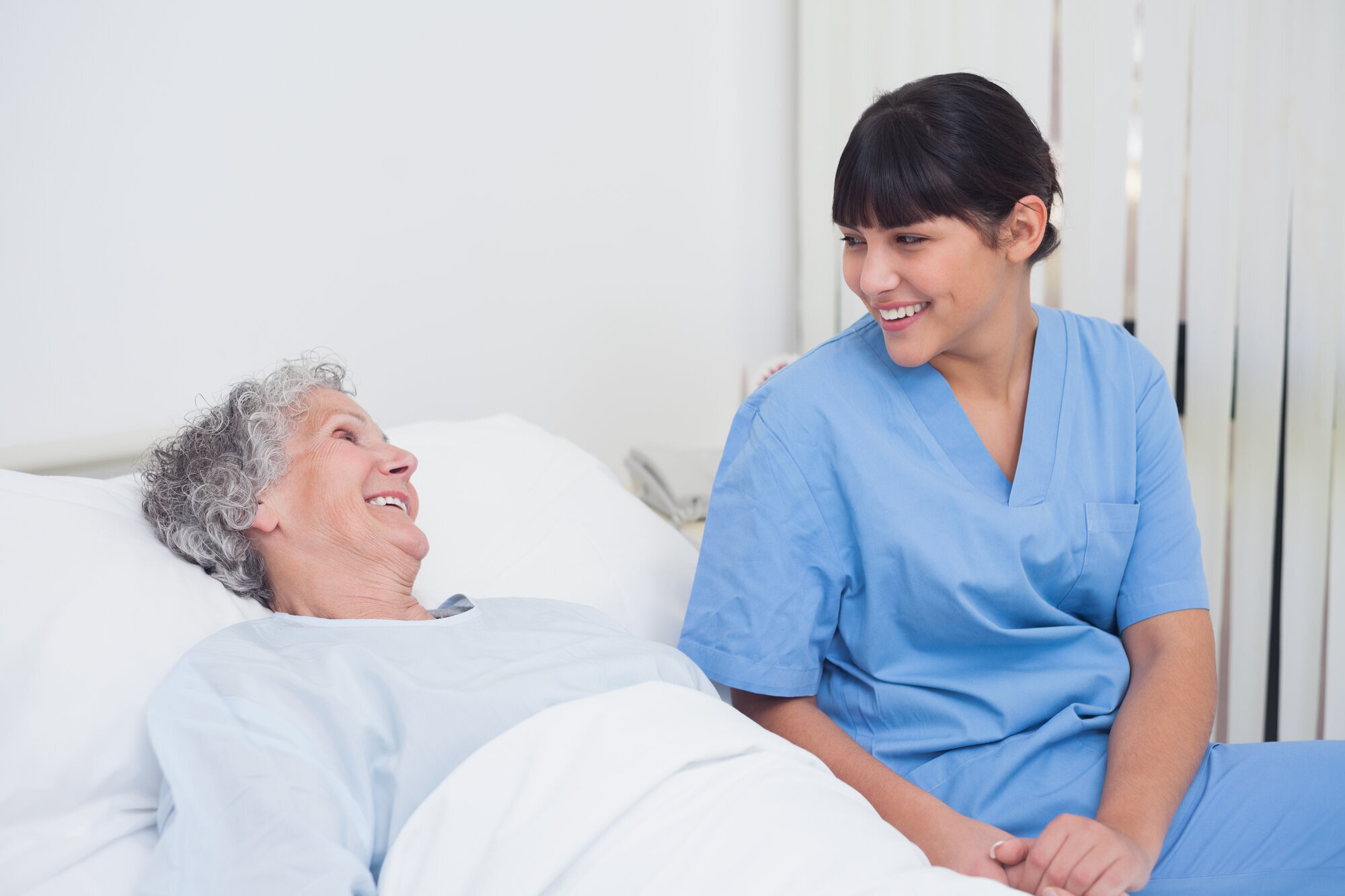 Smil Bildet viser en eldre kvinne som ligger i en seng og smiler til et yngre kvinnelig helsepersonell som smiler tilbake