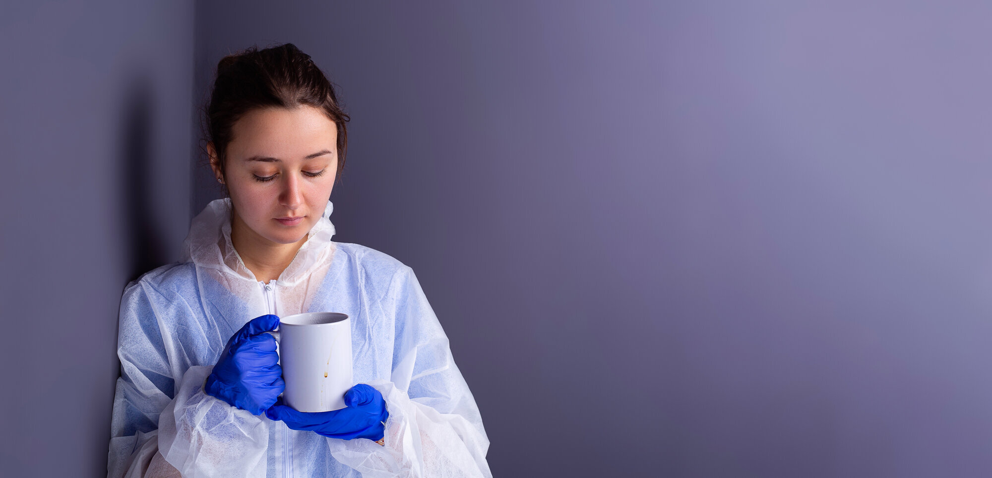 Pleier Bildet viser et ungt, kvinnelig helsepersonell i verneutstyr som ser sliten ut og har en kopp i hånden