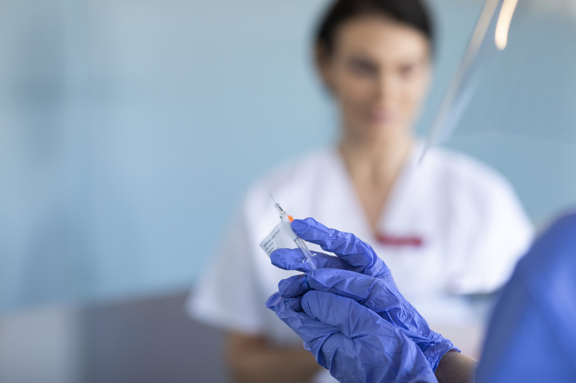 Vaksine, korona Bildet viser en hånd som holder en sprøyte, og en kvinne i bakgrunnen.