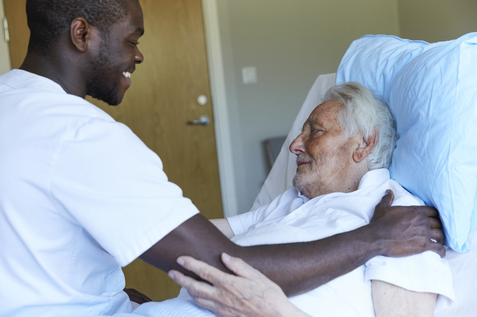 Sykepleier og pasient Bildet viser en mannlig sykepleier som smiler og holder hånden sin på skulderen til en liggende pasient