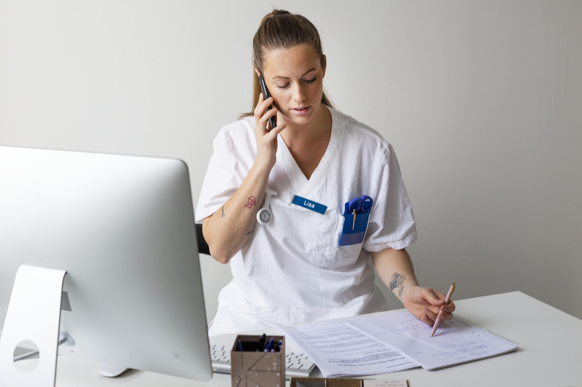 Sykepleier Bildet viser en ung, kvinnelig sykepleier som snakker i telefonen og tar notater.