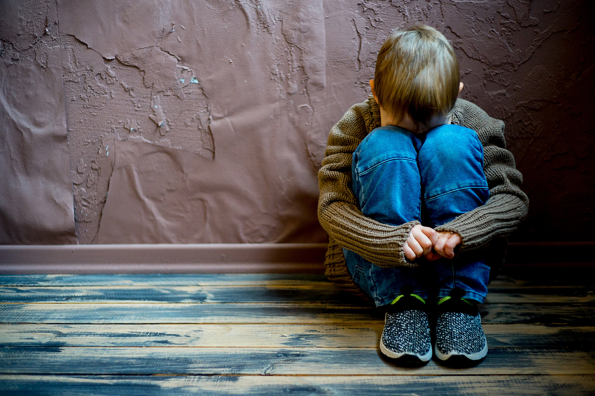 Barn Bildet viser et lite barn som sitter mot en vegg med opptrukne knær og skjuler ansiktet sitt.