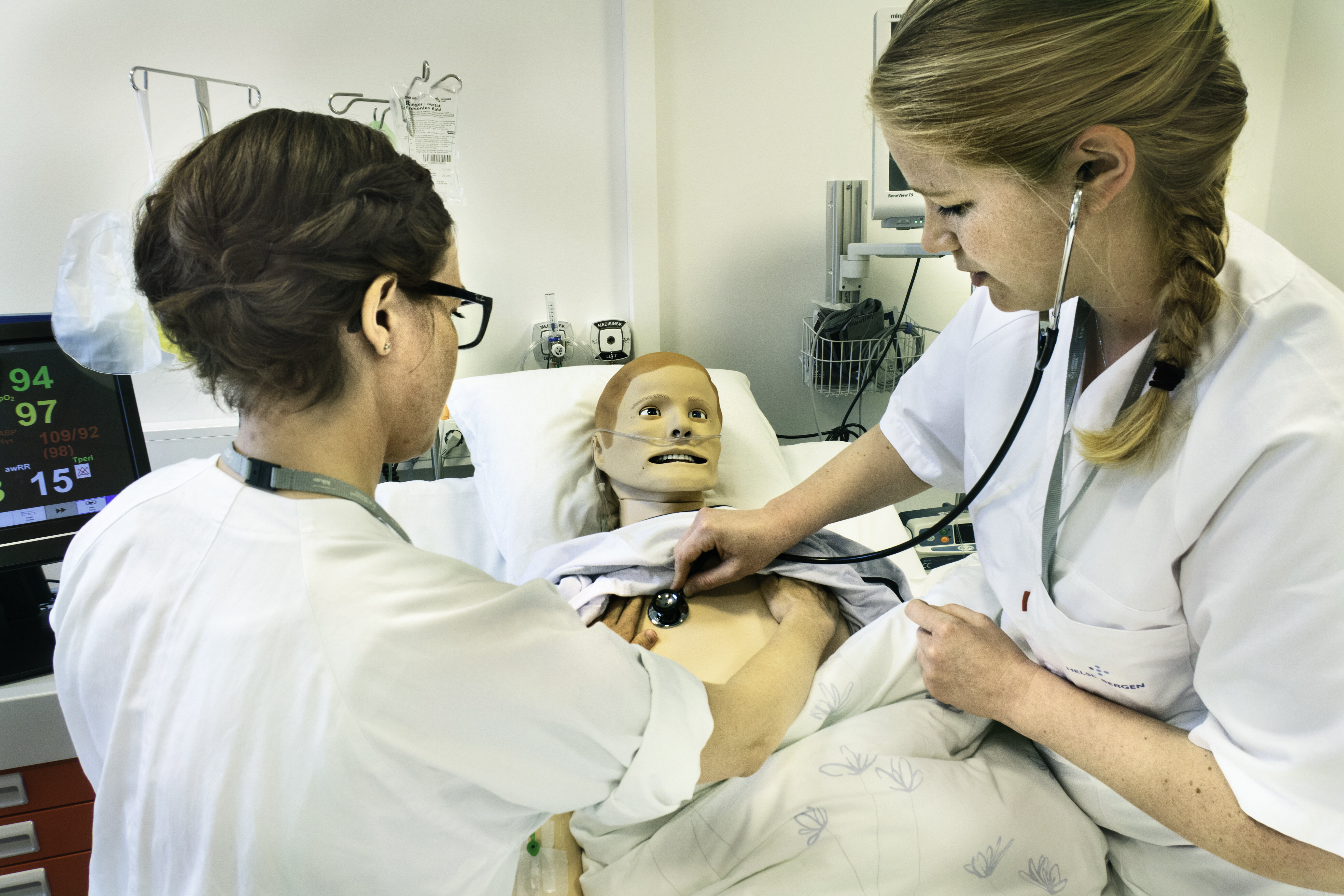 Bildet viser to sykepleiere som står ved en seng. I senga ligger en simuleringsdukke. Den ene sykepleieren lytter etter hjerteslag på dukken