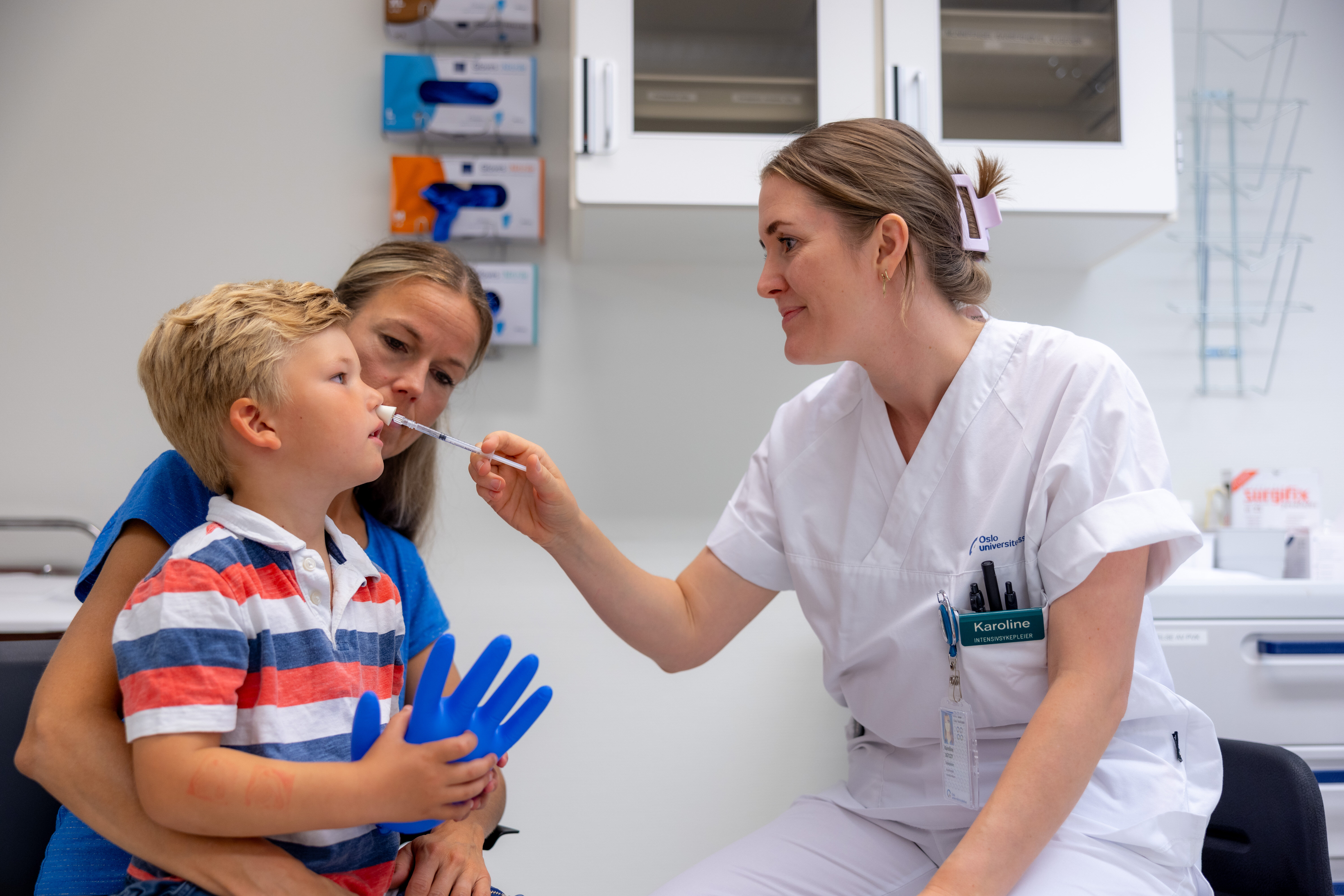 Nasal smertelindring til barn Bildet viser en liten gutt på fanget til sin mor. En sykepleier gir ham nesespray inn i det ene neseboret