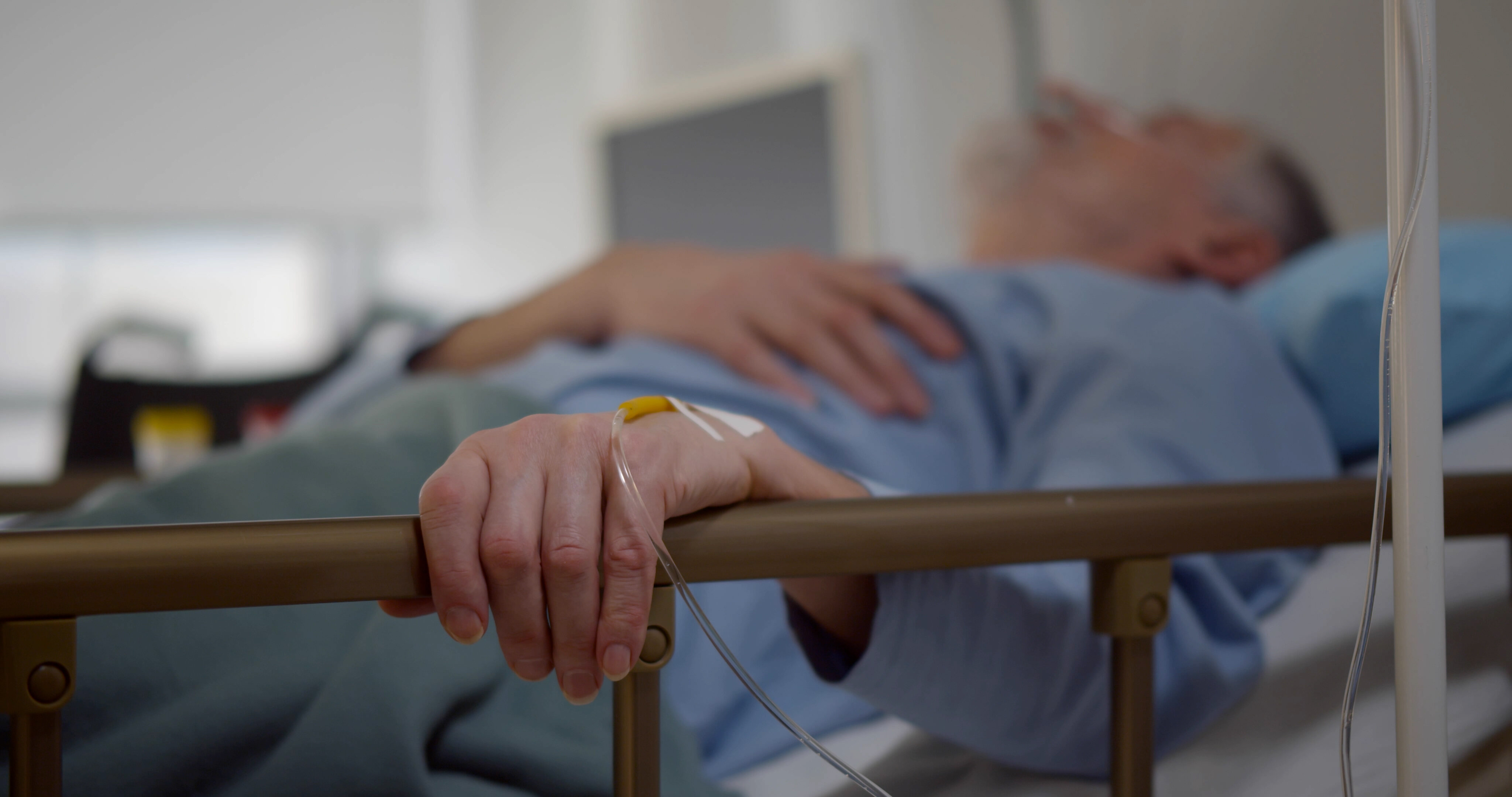 Older man receives intravenous fluid. The photo shows an older man lying in a hospital bed. His left hand is resting on the bed rail. In his hand is a venous access port.