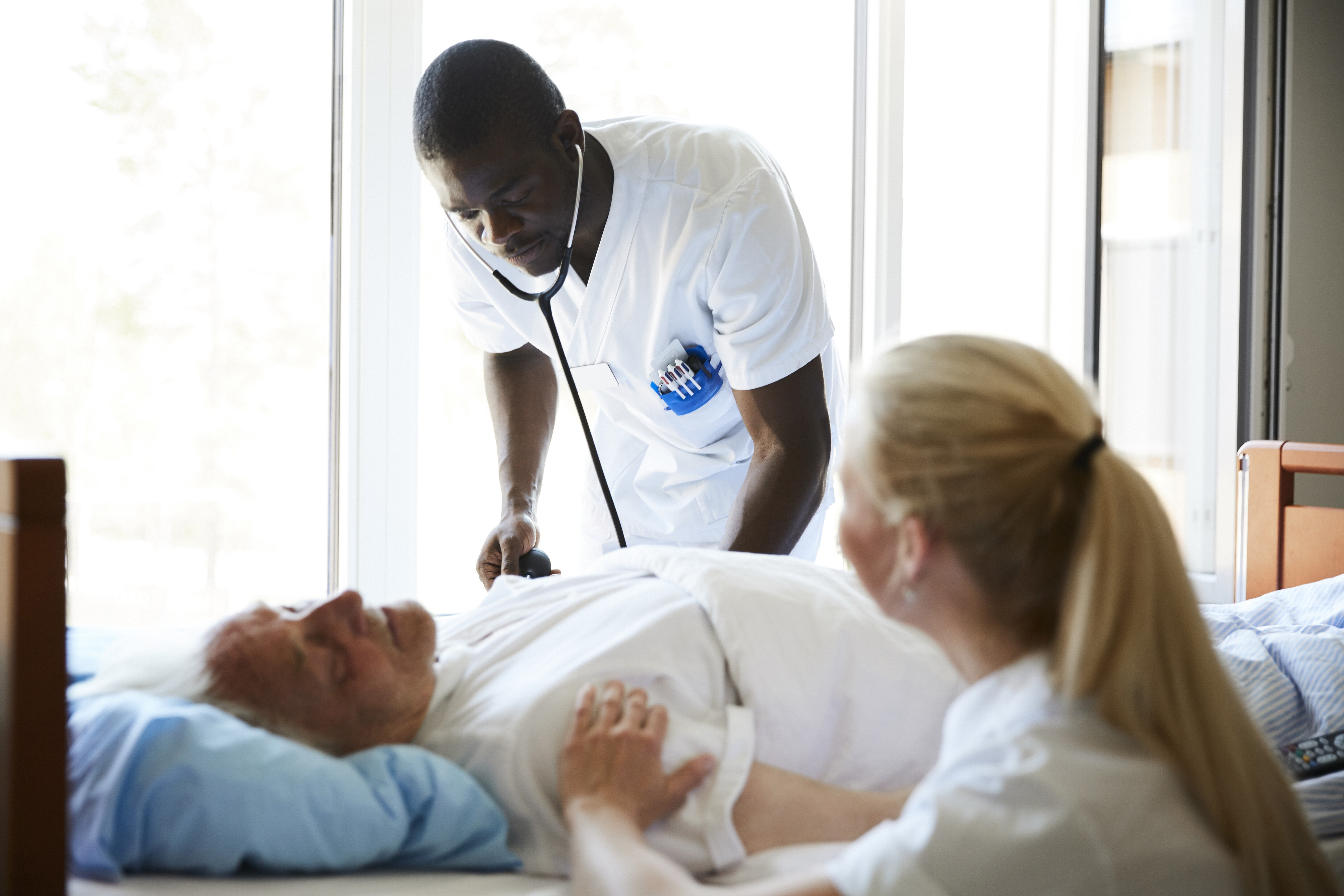Nurses and patient in MABU The photo shows an older patient lying in a hospital bed. A male nurse is bending towards him listening with a stethoscope. A female nurse is sitting beside the bed.