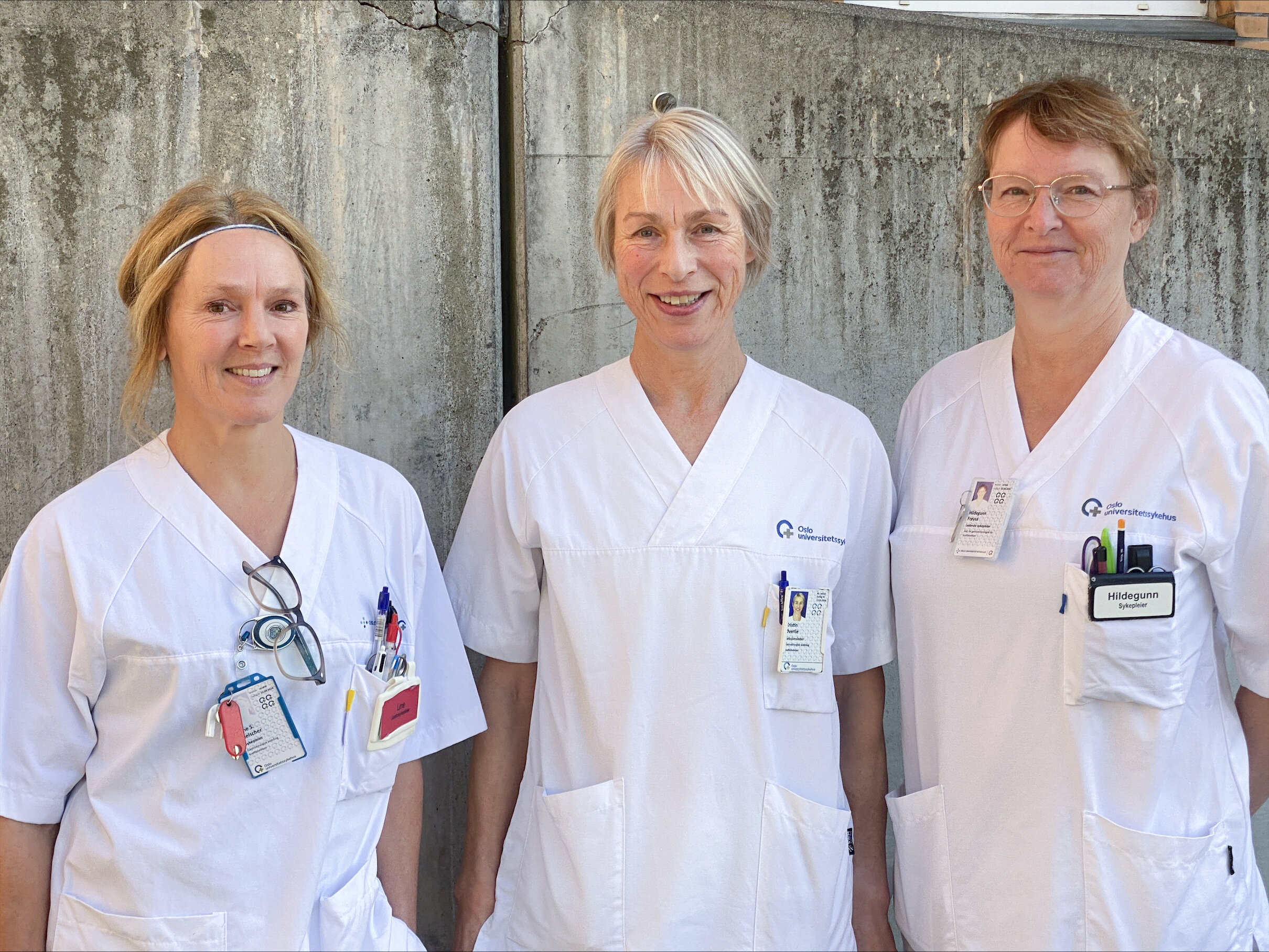 tre sykepleiere står utenfor sykehuset med en sementvegg bak.