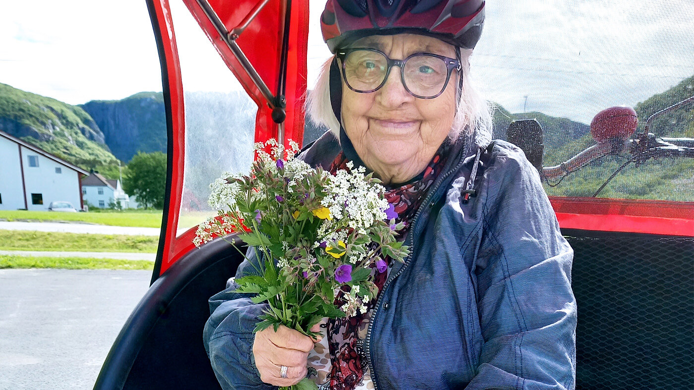 Bildet viser en eldre som sitter i en sykkeltilhenger ute i naturen. I hånden holder hun en bukett markblomster.