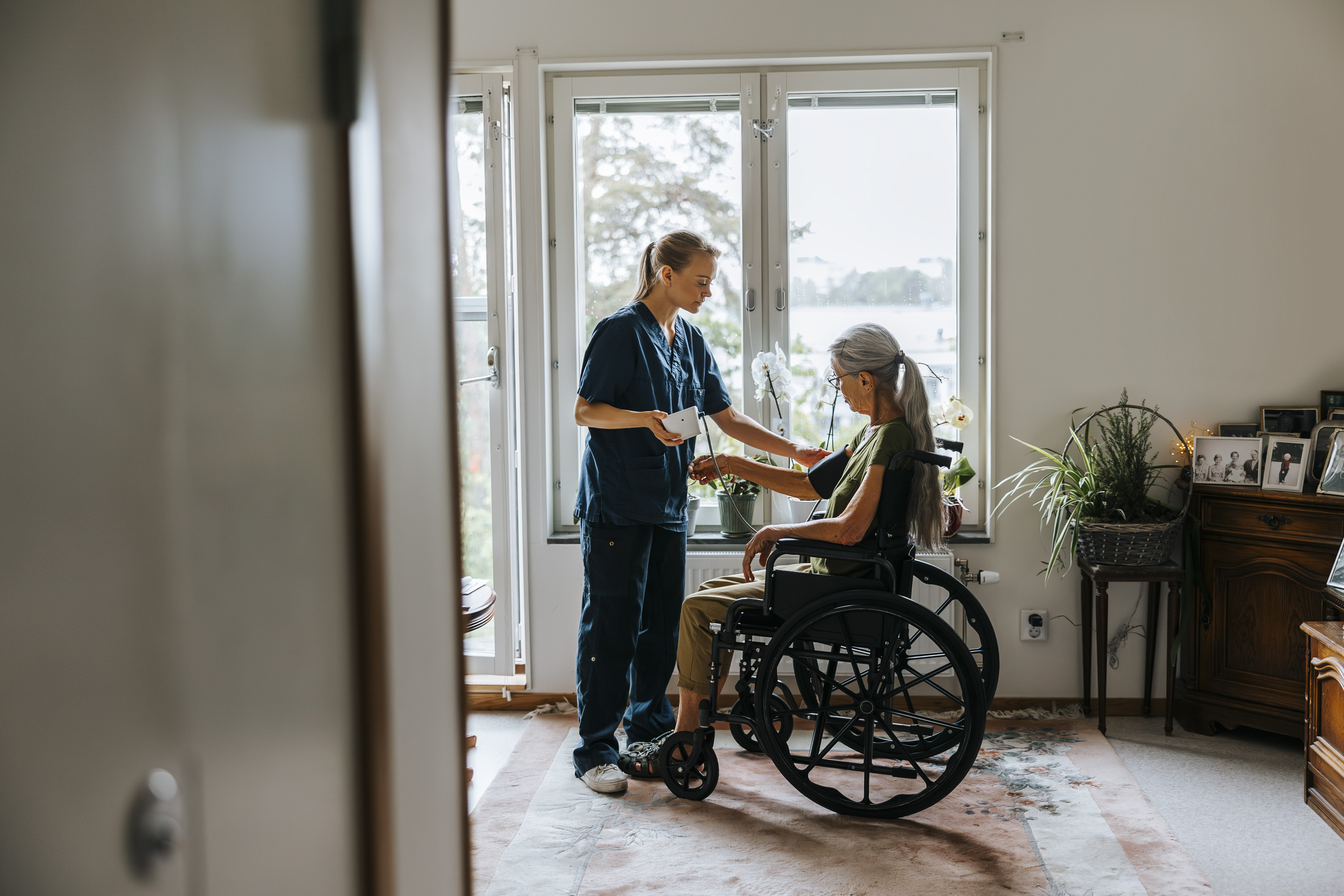 Home care The phowo shows a nurse standing in front of a woman in a wheel chair. The nurse is monitoring her blood pressure. The are in front of a window in the patient's living room