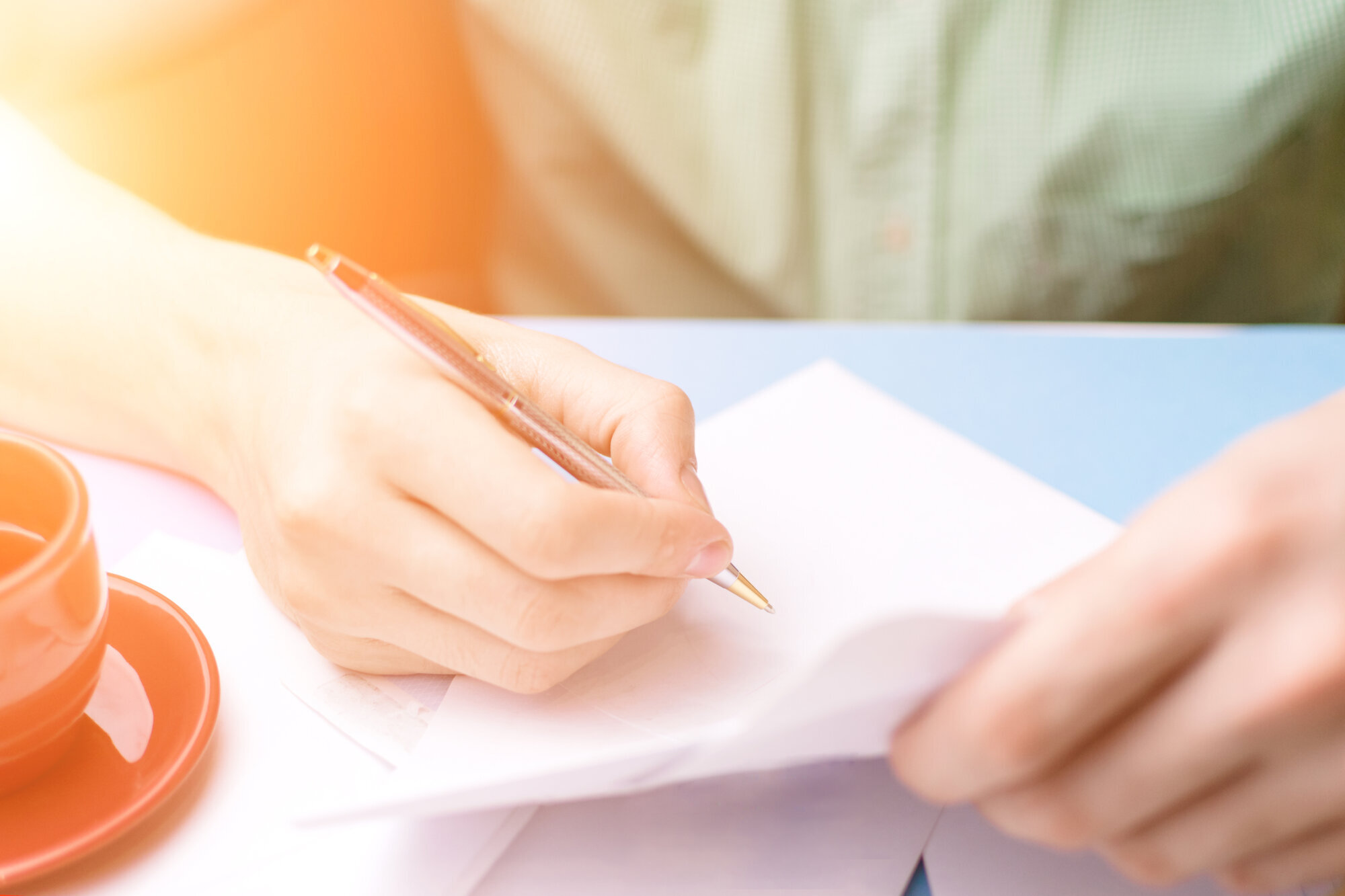 Bildet viser et utsnitt av et menneske som sitter og skriver. Vedkommende har en tekopp ved siden av seg, og solen skinner inn i værelset.