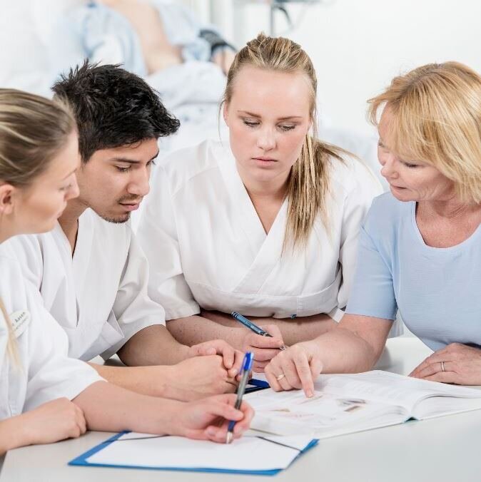 Fasilitator og studenter Fasilitator og tre studenter sitter tett sammen rundt et bord. To av dem holder i en penn, og det er papirark på bordet.