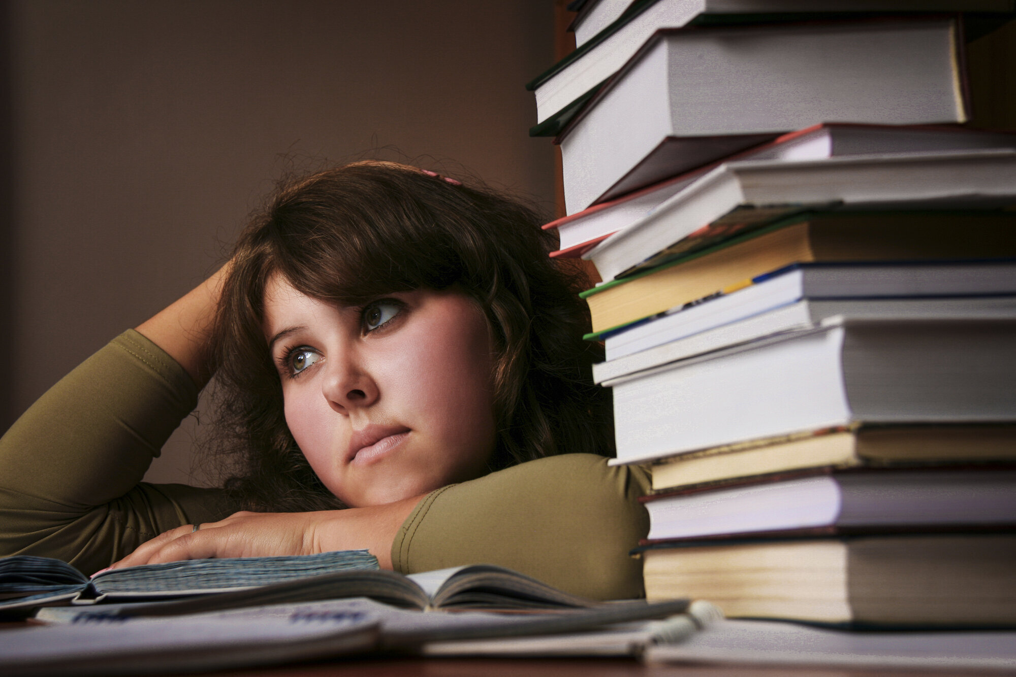 Student med lærebøker Bildet viser en student med et litt oppgitt uttrykk, som ligger over en oppslått bok på pulten. Det ligger en stabel bøker ved siden av henne.