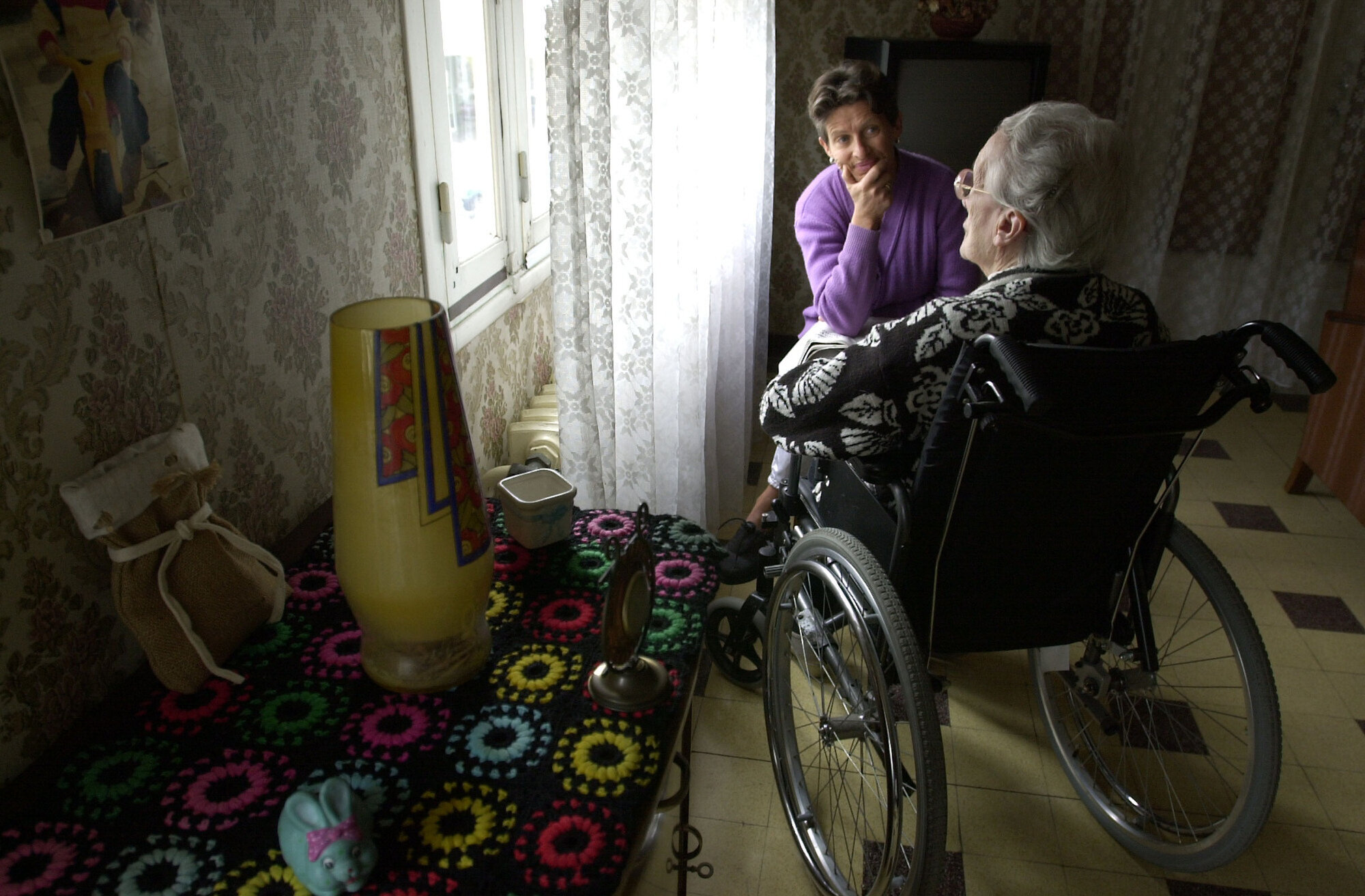 Hjemmesykepleie, eldreomsorg Bildet viser en eldre dame i rullestol sett bakfra og en yngre dame som sitter og lytter til henne.