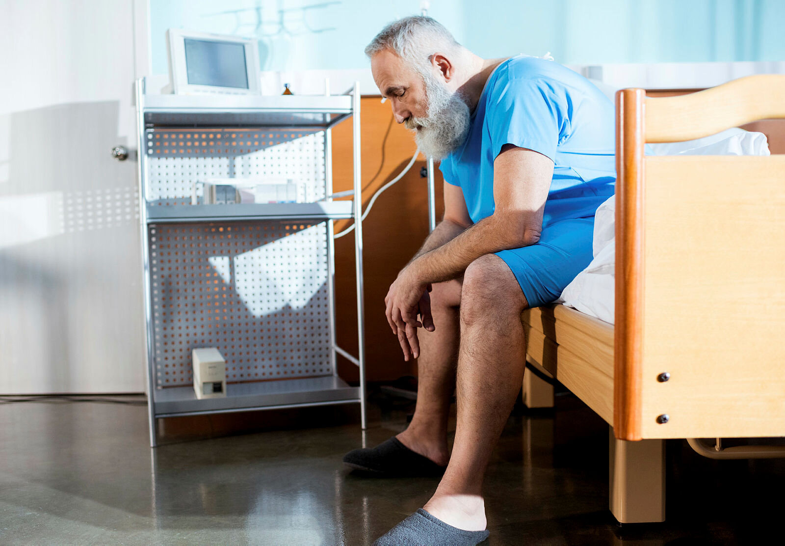 Mann på sykehus Bildet viser en eldre mann med skjegg som sitter på kanten av en sykehusseng med hendene i fanget.