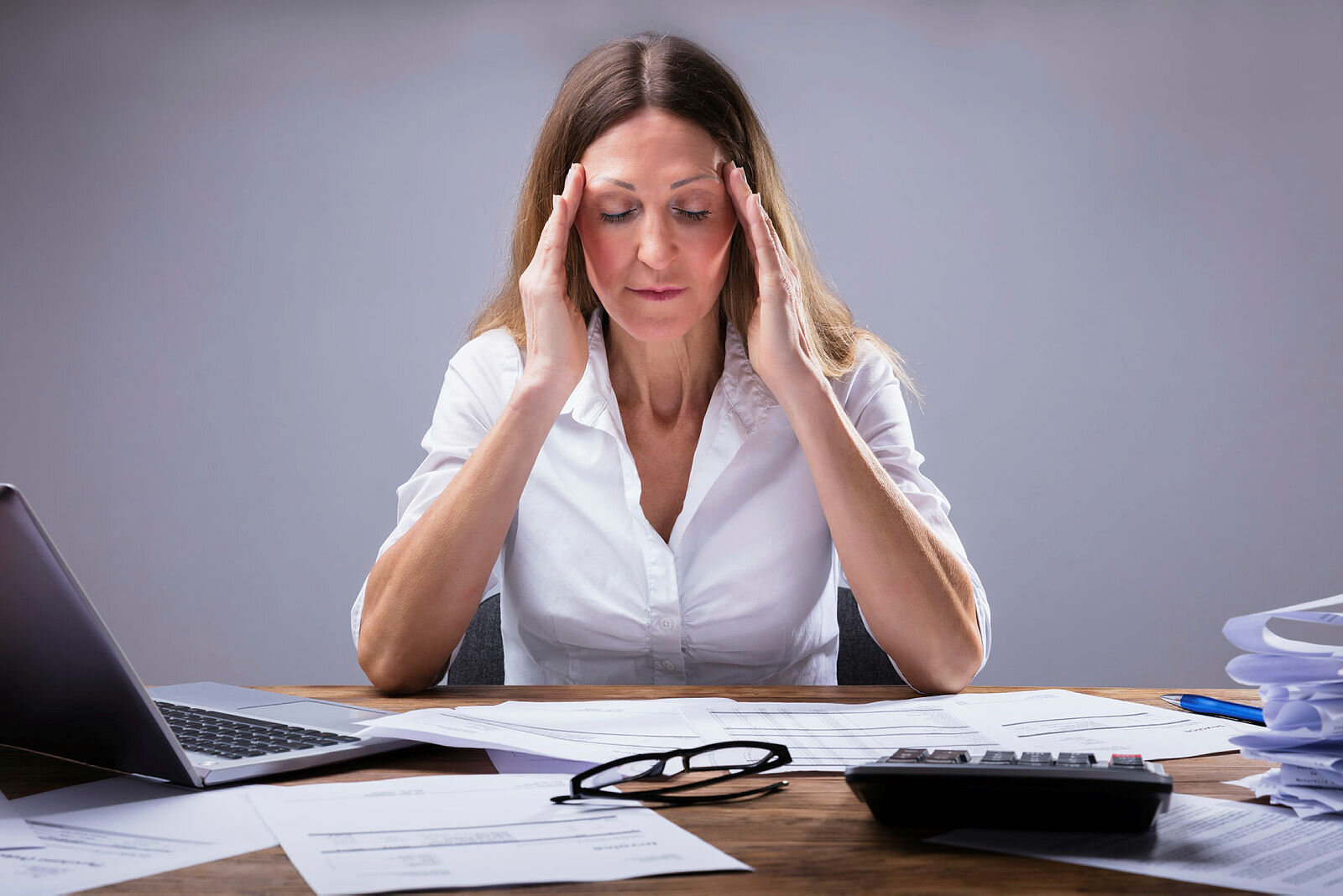 Sliten leder Bildet viser en kvinne som sitter foran et skrivebord med forskjellige permer og rekvisita på og holder seg til tinningene