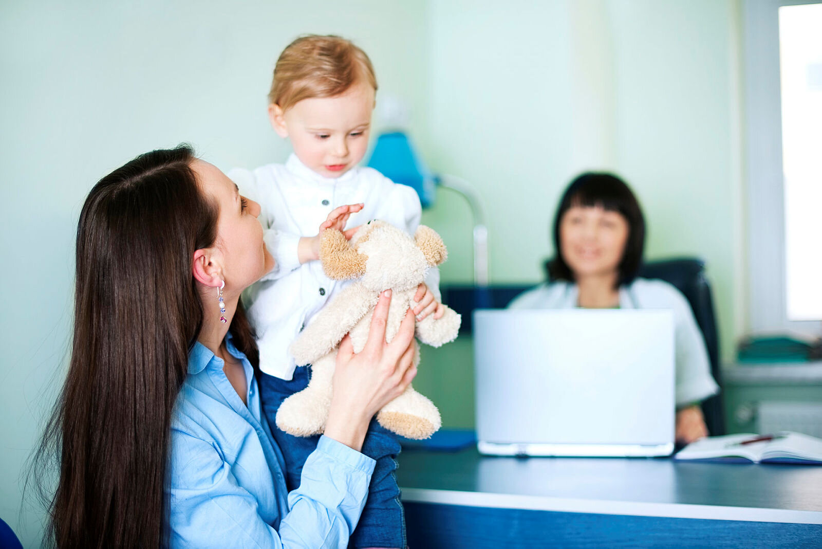 Bildet viser en mor som løfter sitt barn. De sitter foran pulten til helsesøsteren, som skimtes i bakgrunnen.