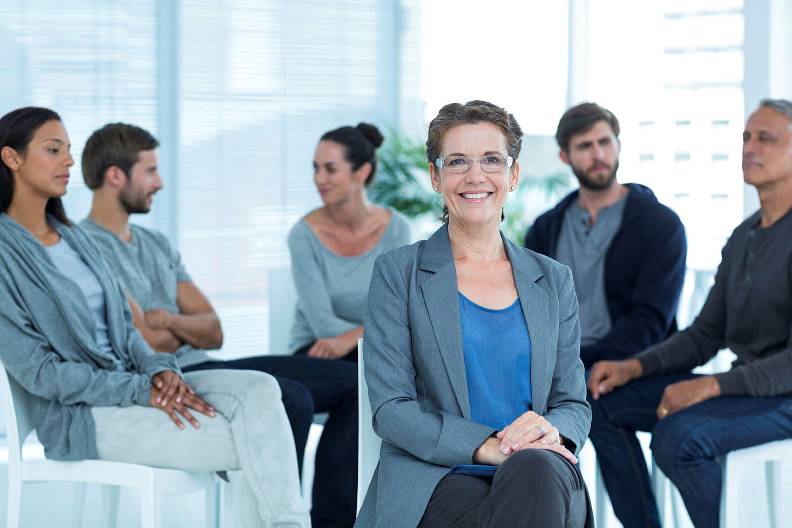 Gruppe Bildet viser en gruppe som sitter i en halvsirkel og en kvinne som sitter med ryggen til gruppen og ser på oss