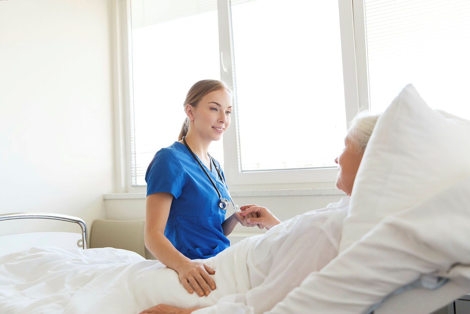 Sykepleier sitter på sengekanten til eldre, sengeliggende kvinnelig pasient.