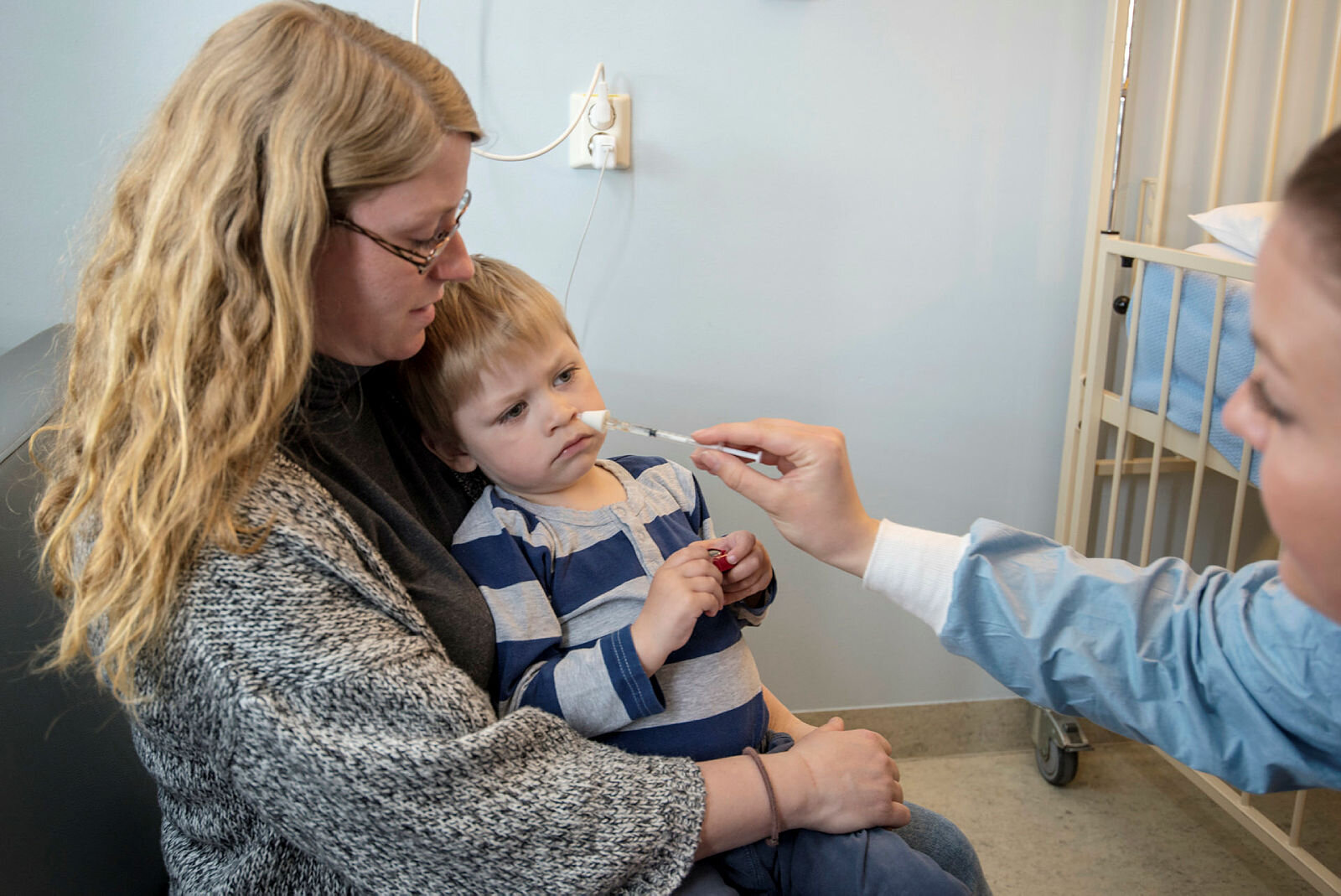 Oliver får nesespray Bildet viser Oliver som sitter på mammas fang og får nesespray av en sykepleier.