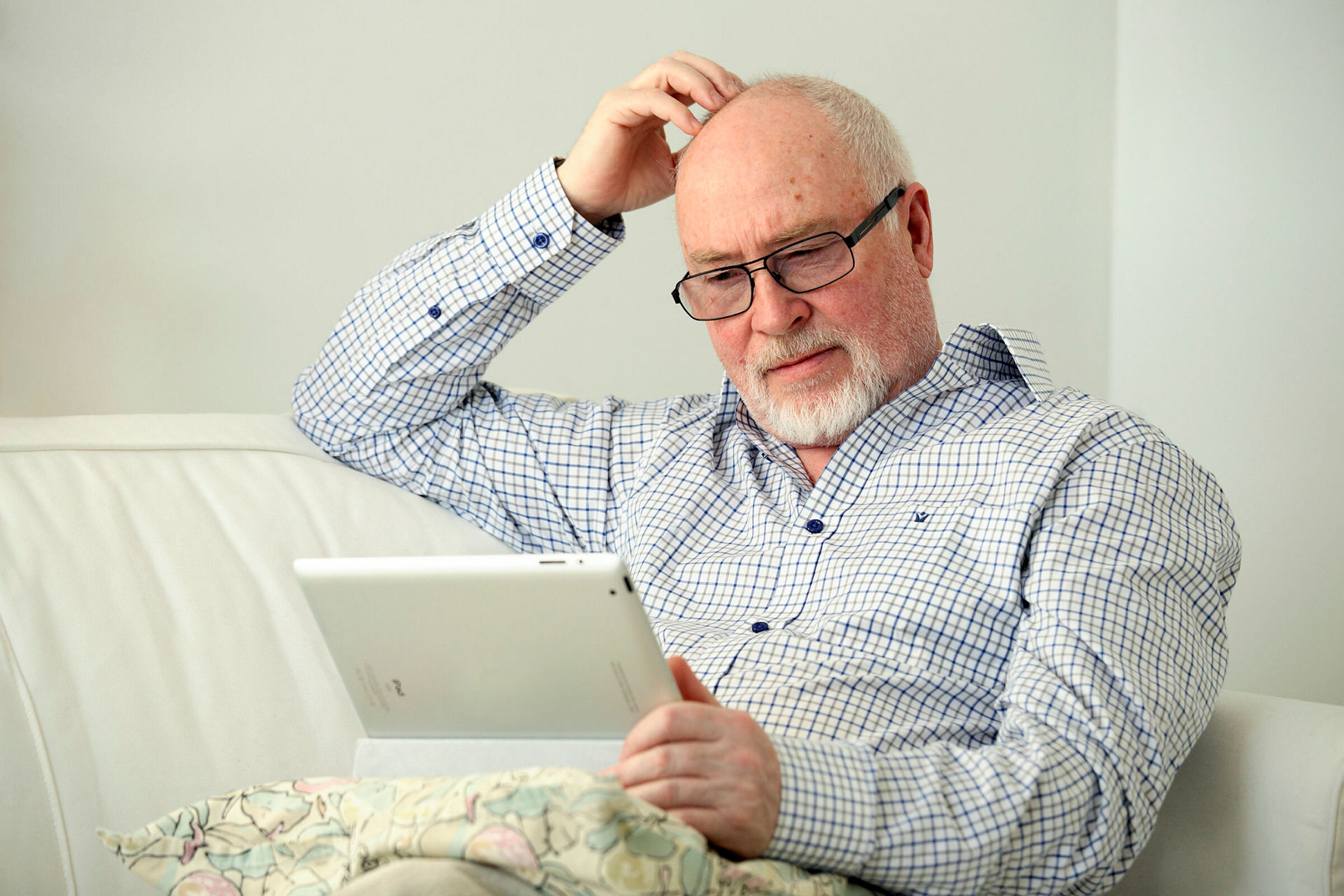 Mann med brett Bildet viser en eldre mann som sitter i en sofa med et nettbrett på fanget og klør seg litt i hodet