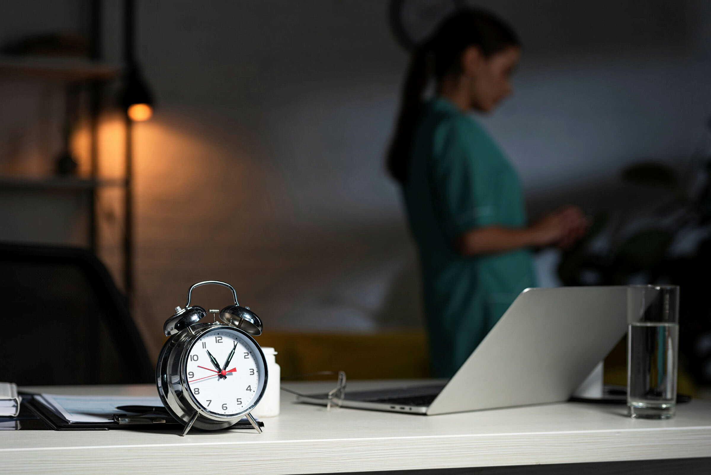 Sykepleier på nattevakt Bildet viser en vekkerklokke i forgrunnen og en sykepleier og en tent lampe i bakgrunnen. Rommet er ganske mørkt.