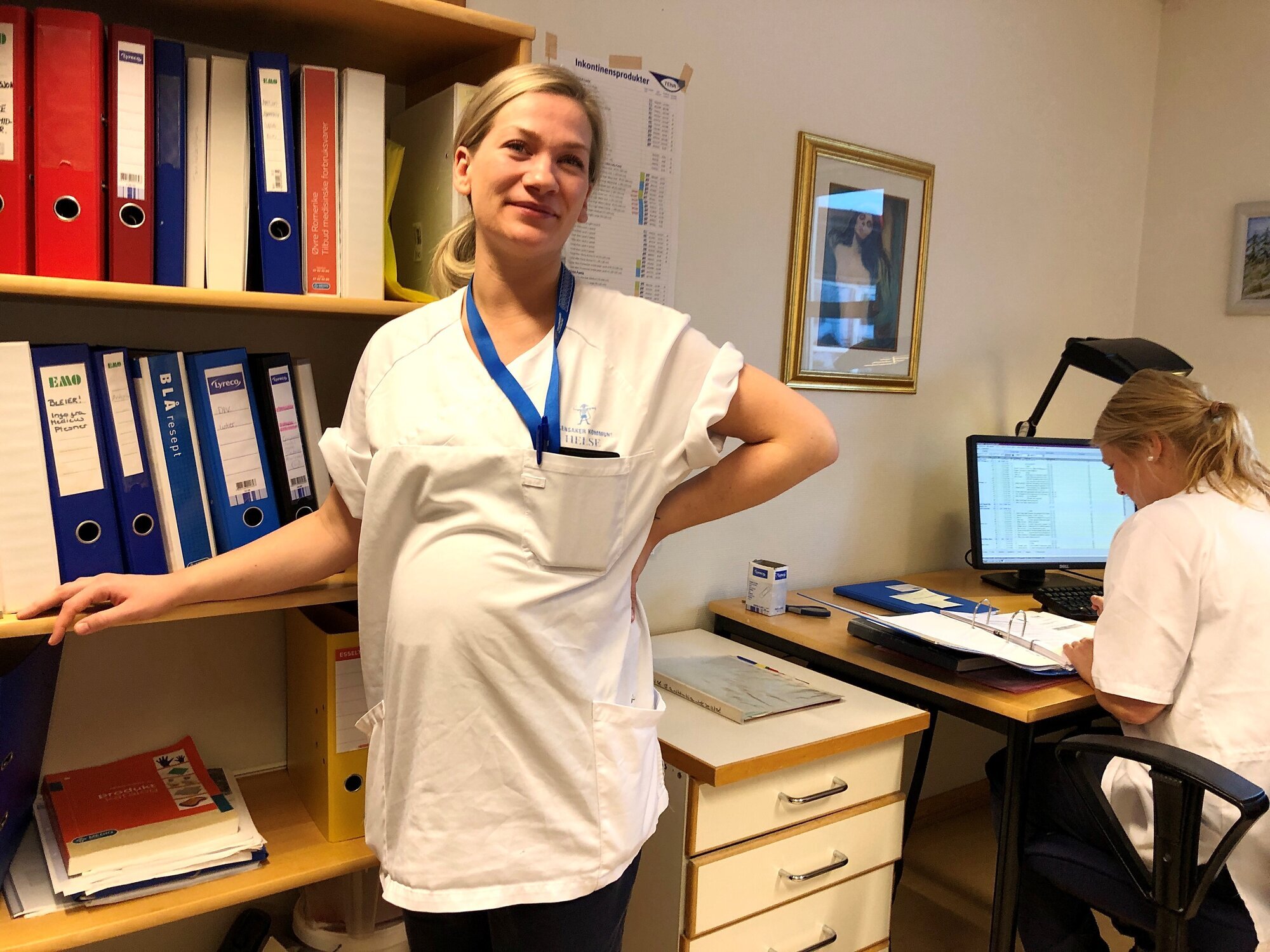 Høygravid sykepleier tar seg til ryggen og lener seg til bokhylle. Kollega sitter foran skjerm ved sine av henne.