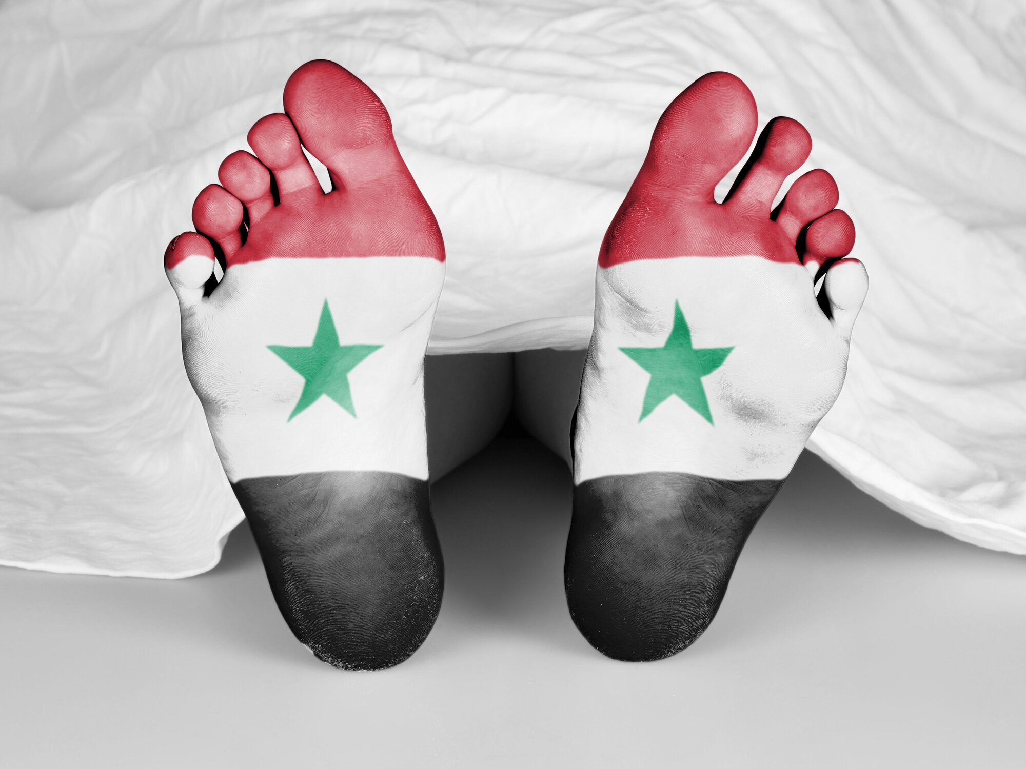 En død kropp under et laken hvor beina stikker frem. Det er tegnet det Syriske flagget på fotsålene. En død kropp under et laken hvor beina stikker frem. Det er tegnet det Syriske flagget på fotsålene.