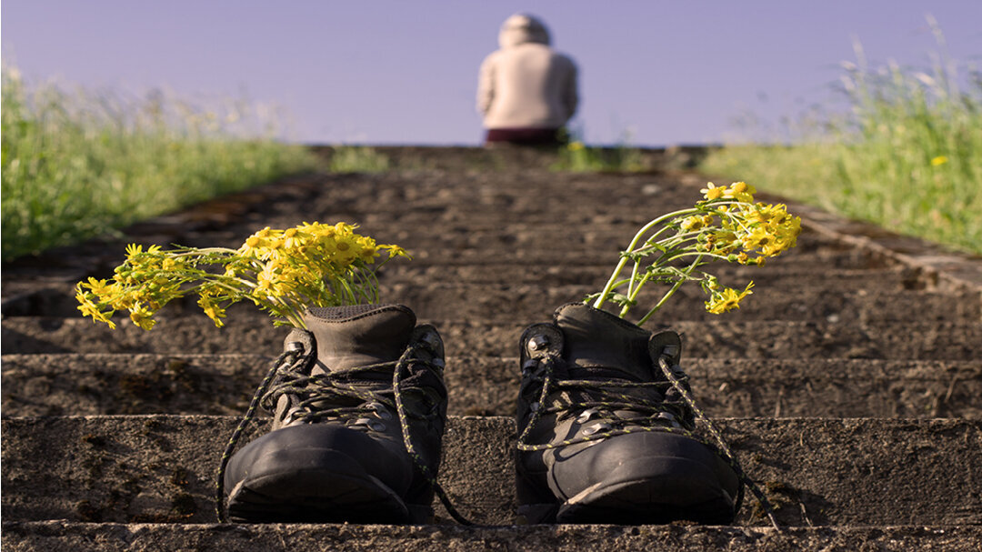Et menneske sitter på trappen. Bildet viser et menneske som sitter med ryggen til øverst i en trapp ute i det fri. Det står et par sko for seg selv på trappen med blomster i.