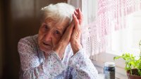 Bildet viser en trist, gammel dame som lener hodet mot hendene sine. Hun sitter ved kjøkkenbordet med et pilleglass foran seg.