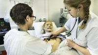 Bildet viser to sykepleiere som står ved en seng. I senga ligger en simuleringsdukke. Den ene sykepleieren lytter etter hjerteslag på dukken