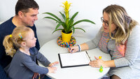 Parent contact in the school health service The photo shows a father with his daughter on his lap. They are sitting at a table opposite a woman leaning towards them. She has a notebook in front of her.
