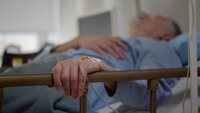 Older man receives intravenous fluid. The photo shows an older man lying in a hospital bed. His left hand is resting on the bed rail. In his hand is a venous access port.