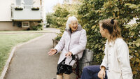 Bildet viser en smilende kvinne med rullator som snakker med en pleier ute