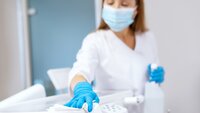 Smittehåndtering Bildet viser en sykepleier med munnbind og hansker. Hun holder en sprayflaske i hånden og tørker med en fille