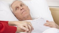 Eldre mann i sykehusseng Bildet viser en syk, eldre mann som ligger i en sykehusseng. En dames hender holder hans.