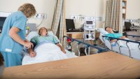 Sykepleier undersøker pasient. En sykepleier undersøker en pasient på sengepost. En annen pasient ligger i sengen ved siden av.