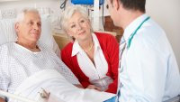 Mann på sykehuset med besøk av kona og legen Bildet viser en eldre mann som ligger i senga på sykehuset. Kona sitter ved siden av, og legen er på visitt.